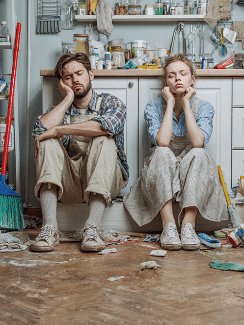 Aide ménagère à domicile – Major'Dom 67 Couple assis par terre dans une cuisine en désordre, débordé par le ménage, faisant appel à Major'Dom 67